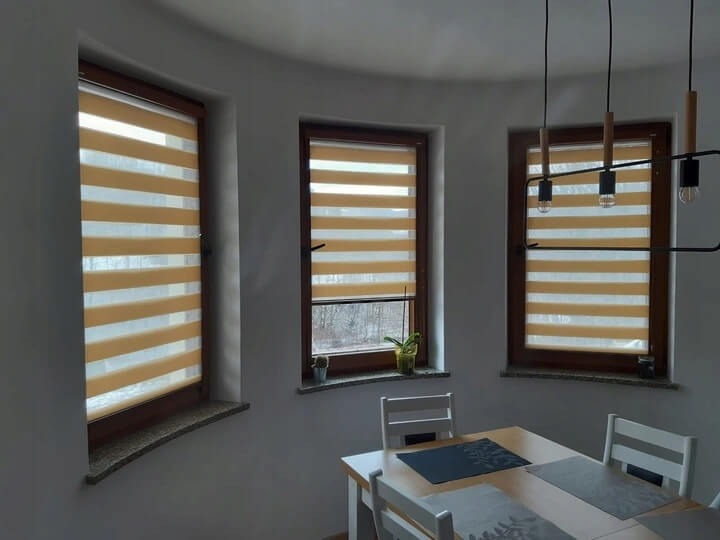 Beżowe rolety dzień noc w drewnianych oknach, widoczne zza stołu w jadalni w Stoczku Łukowskim. Dekoracyjne osłony okienne z poziomymi pasami.