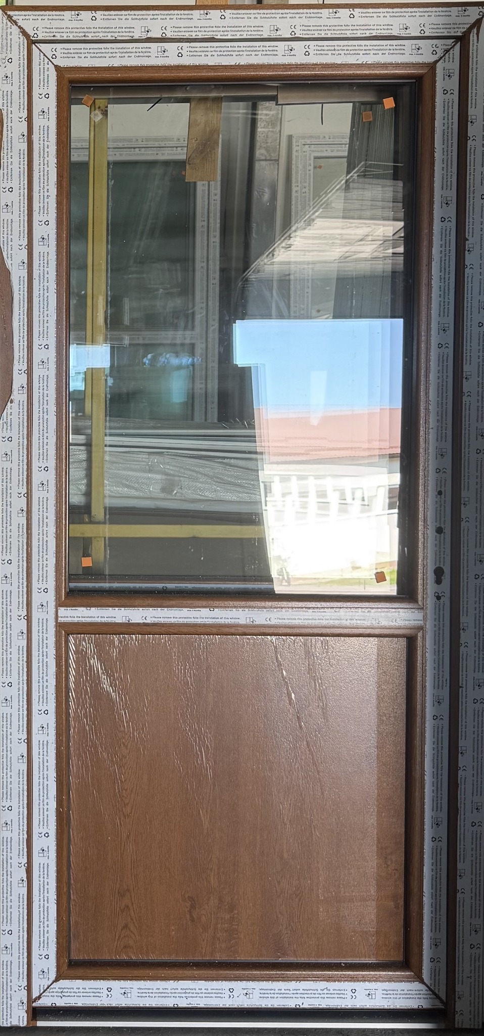 Drewnopodobne okno PCV z szybą, widoczne folie ochronne na ramie. W tle inne okna i elementy konstrukcyjne. Ujęcie z bliska, prezentujące detale wykończenia.