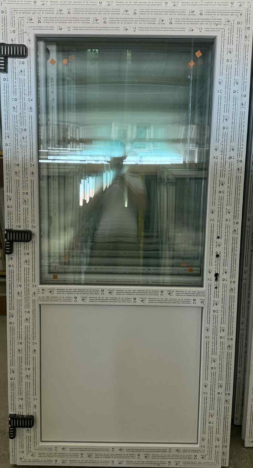 Białe okno PCV z szybą i panelem dolnym, widoczne detale ramy z oznaczeniami. Ujęcie z bliska, prezentujące produkt w jasnym oświetleniu.