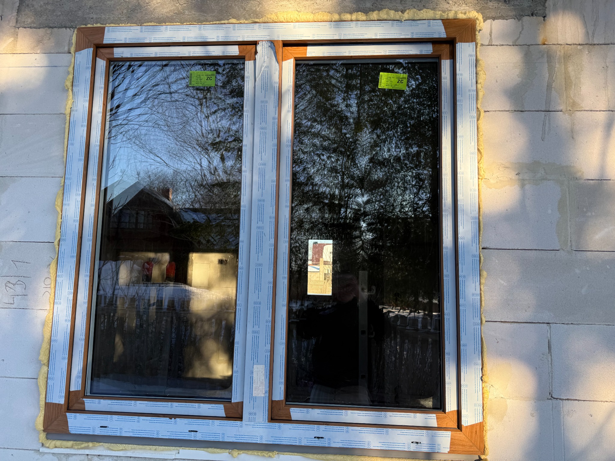 Nowe, dwuskrzydłowe okno z brązową ramą, zamontowane w surowej ścianie z betonu komórkowego. Widoczne pianka montażowa i folia ochronna na profilach.