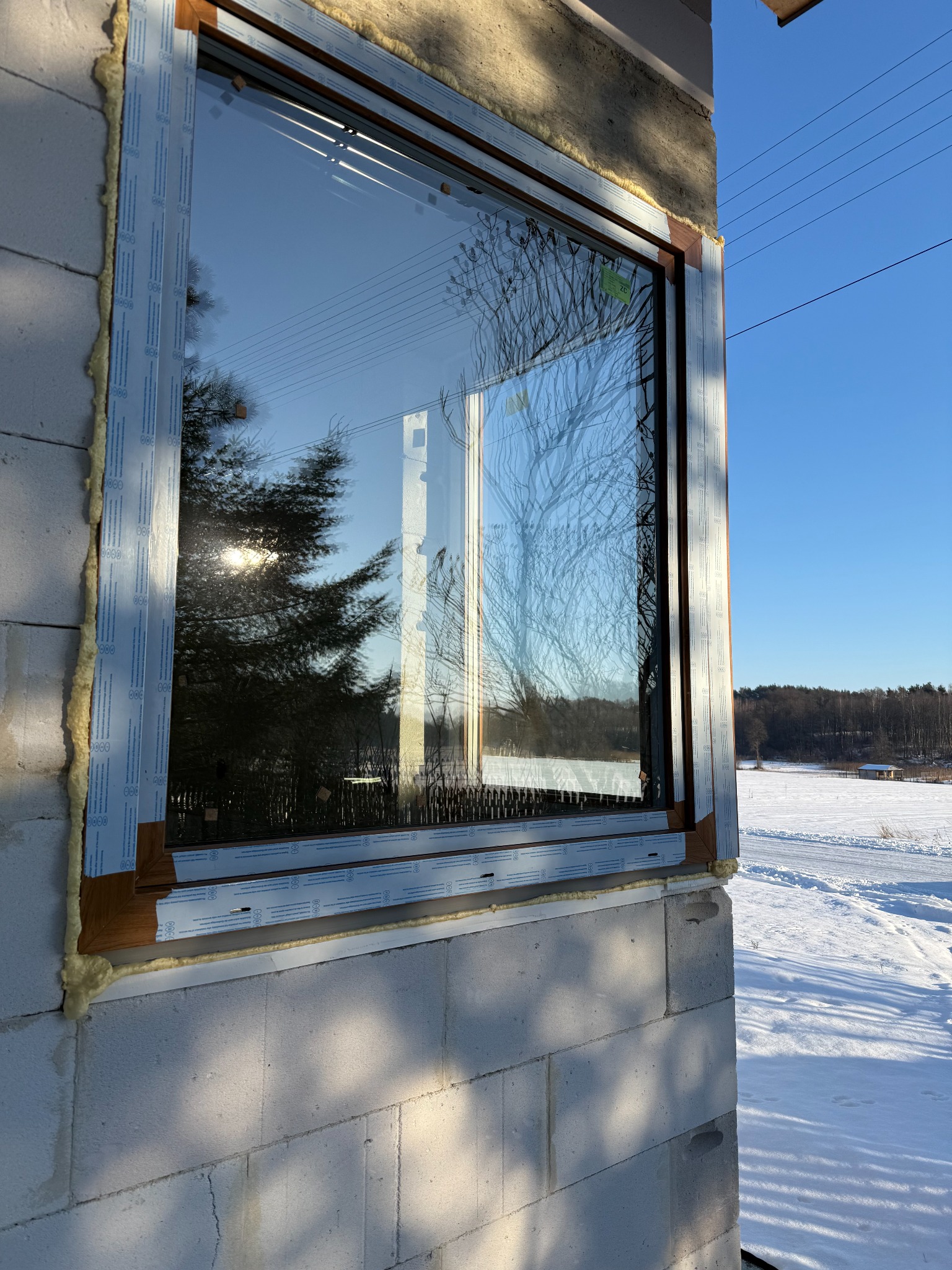 Nowe okno z brązową ramą, zamontowane w surowej ścianie z betonu komórkowego. Widoczne pianka montażowa i folia ochronna. W szybie odbija się zimowy krajobraz.