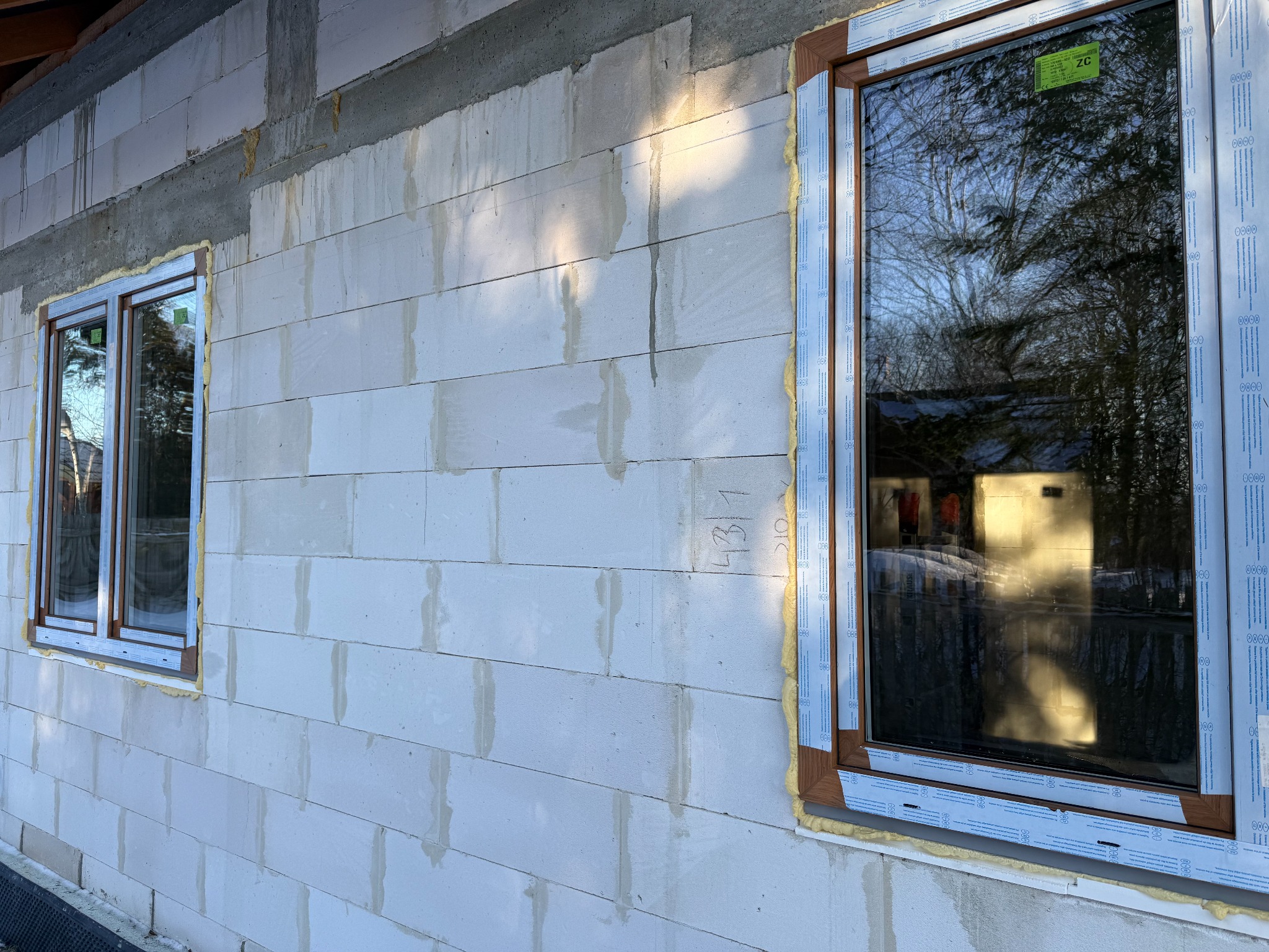 Montaż okien PCV w surowym budynku z betonu komórkowego. Widoczne dwa okna z brązowymi ramami, zabezpieczone folią ochronną i uszczelnione pianką montażową.