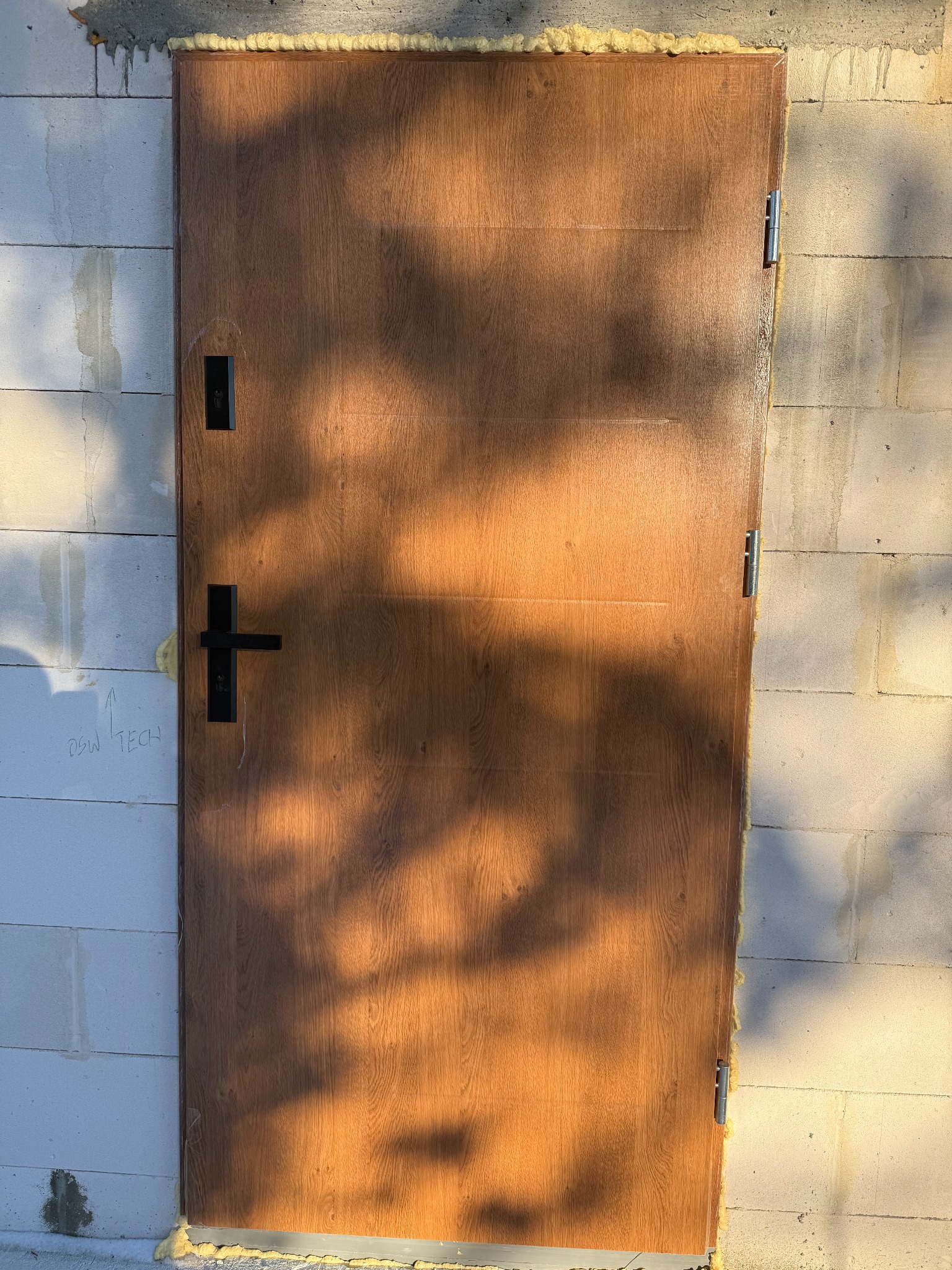 Brązowe drzwi zewnętrzne z czarną klamką, zamontowane w surowej ścianie z betonu komórkowego, uszczelnione pianką montażową. Na drzwiach widoczne cienie drzew.