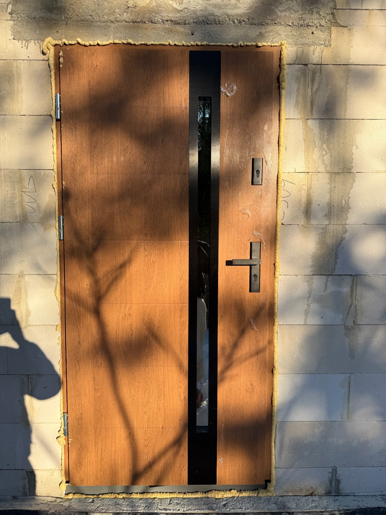 Brązowe drzwi zewnętrzne z czarnym pionowym przeszkleniem, zamontowane w surowej ścianie z betonu komórkowego, uszczelnione pianką montażową. Cień drzewa na drzwiach.
