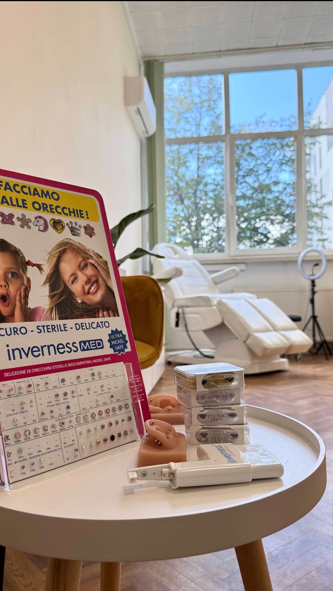 Zestaw do przekłuwania uszu Inverness MED na stoliku w gabinecie, z katalogiem kolczyków i modelem ucha. W tle fotel zabiegowy i lampa pierścieniowa.