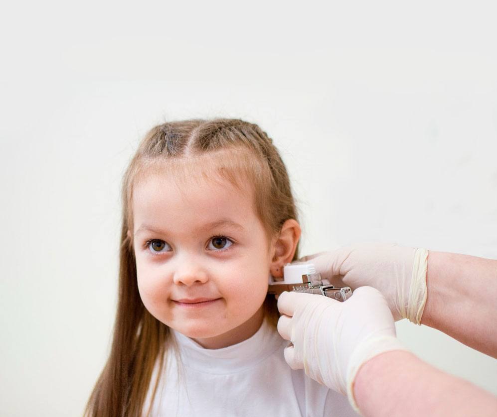 Przekłuwanie ucha u dziecka za pomocą specjalistycznego urządzenia. Drobny kolczyk w uchu, białe rękawiczki ochronne, jasne tło. Delikatny uśmiech dziewczynki.