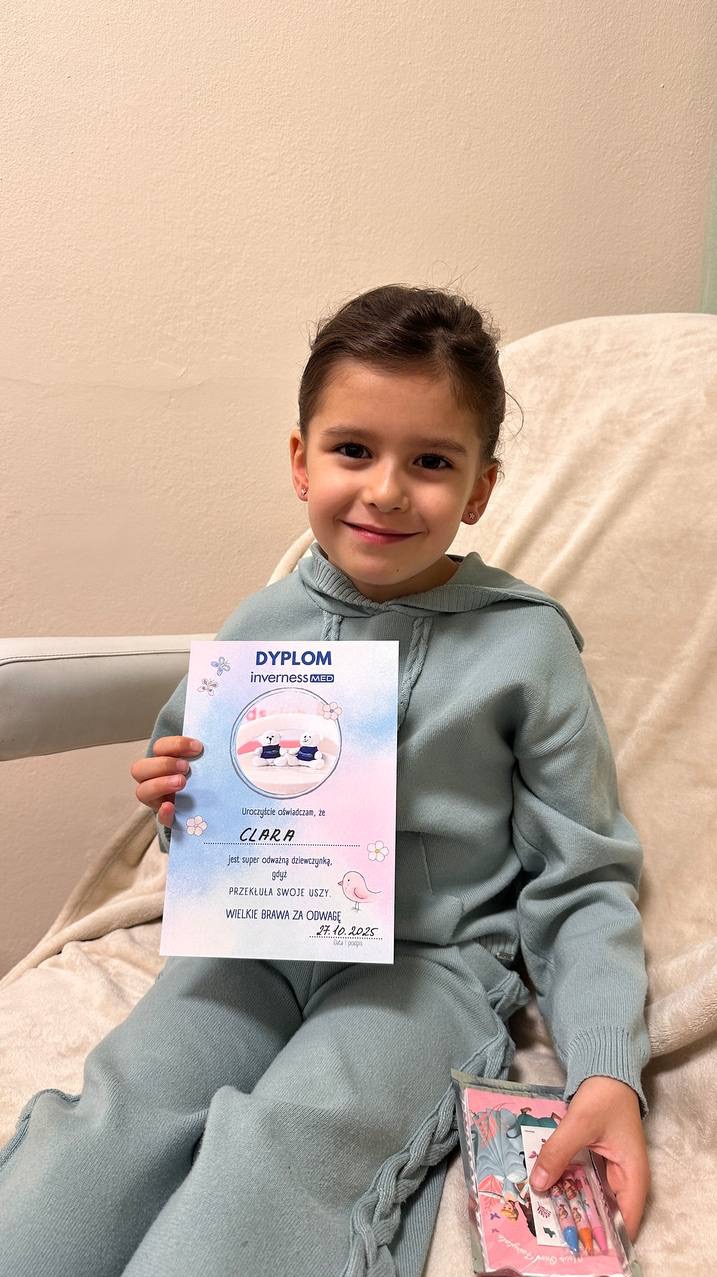 Uśmiechnięta dziewczynka w dresie prezentuje dyplom za przekłucie uszu, trzymając kredki. Jasne tło, naturalne światło. Inverness MED.