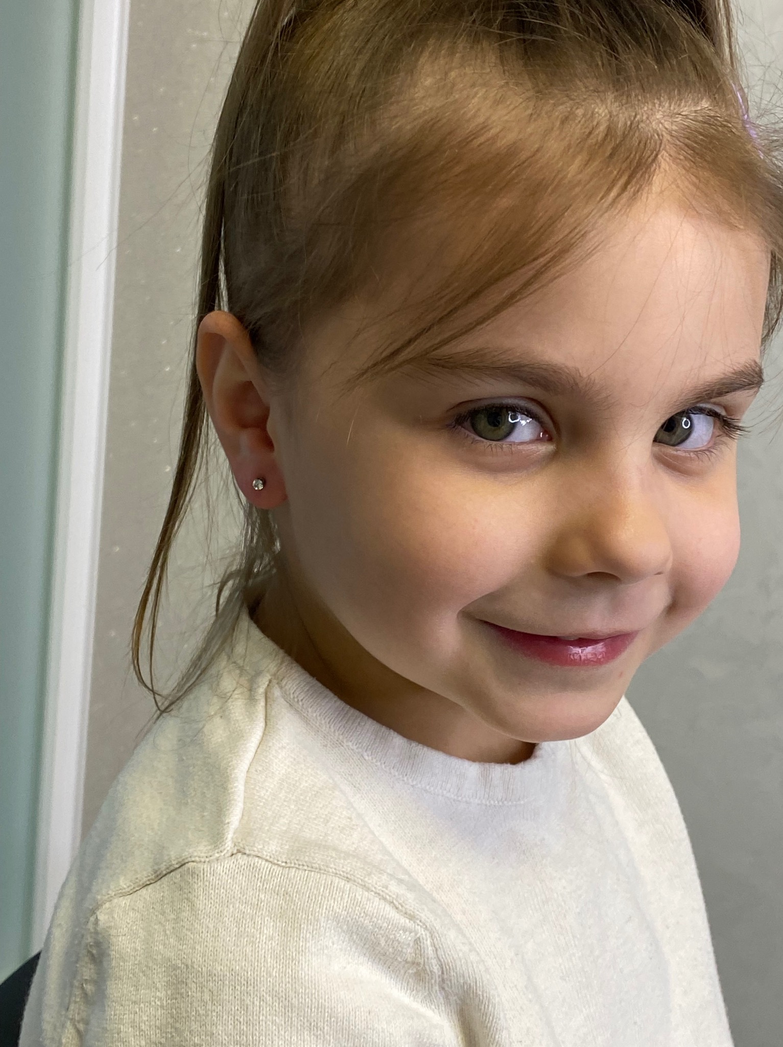 Uśmiechnięta dziewczynka z nowym kolczykiem w uchu, zbliżenie na twarz. Delikatny makijaż ust. Włosy upięte w wysoki kucyk.