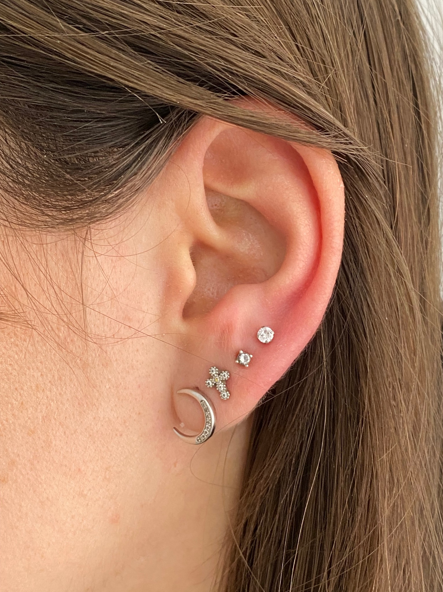 Ucho kobiety z kolczykami: księżyc, krzyż, gwiazda i pojedynczy kamień. Widoczne włosy i fragment policzka. Delikatna biżuteria w srebrnym odcieniu.