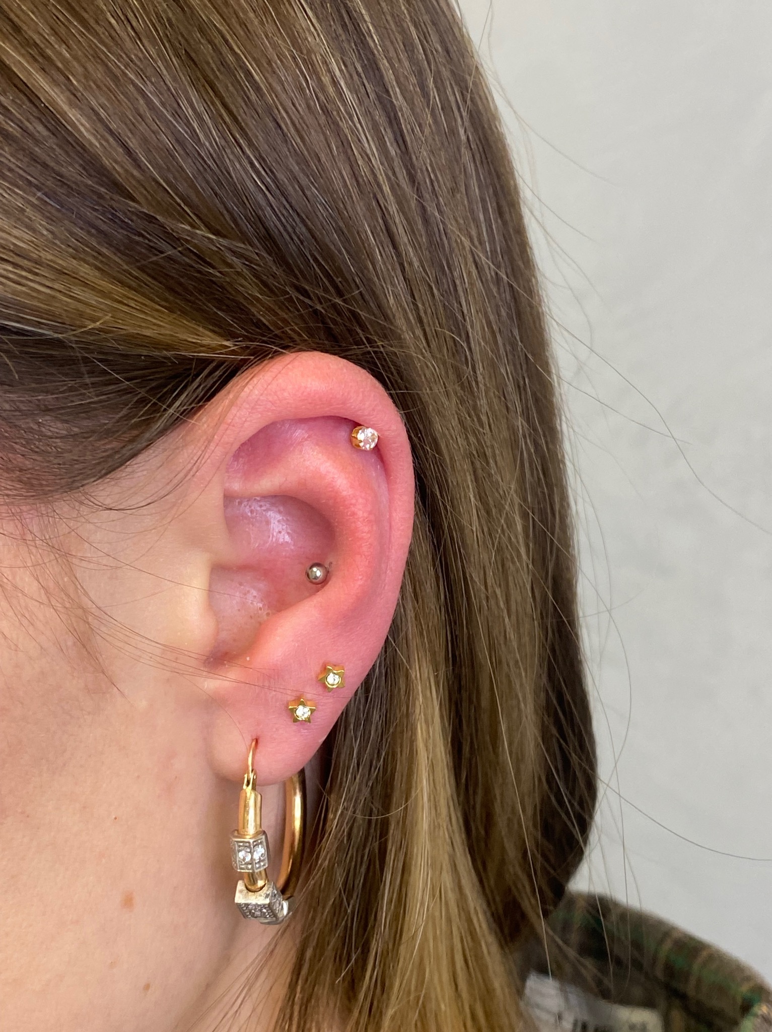 Ucho kobiety z kilkoma kolczykami: helix, conch, lobe. Złote kolczyki w kształcie gwiazdek i kółko z ozdobnymi elementami. Widoczne brązowe włosy i fragment ubrania.