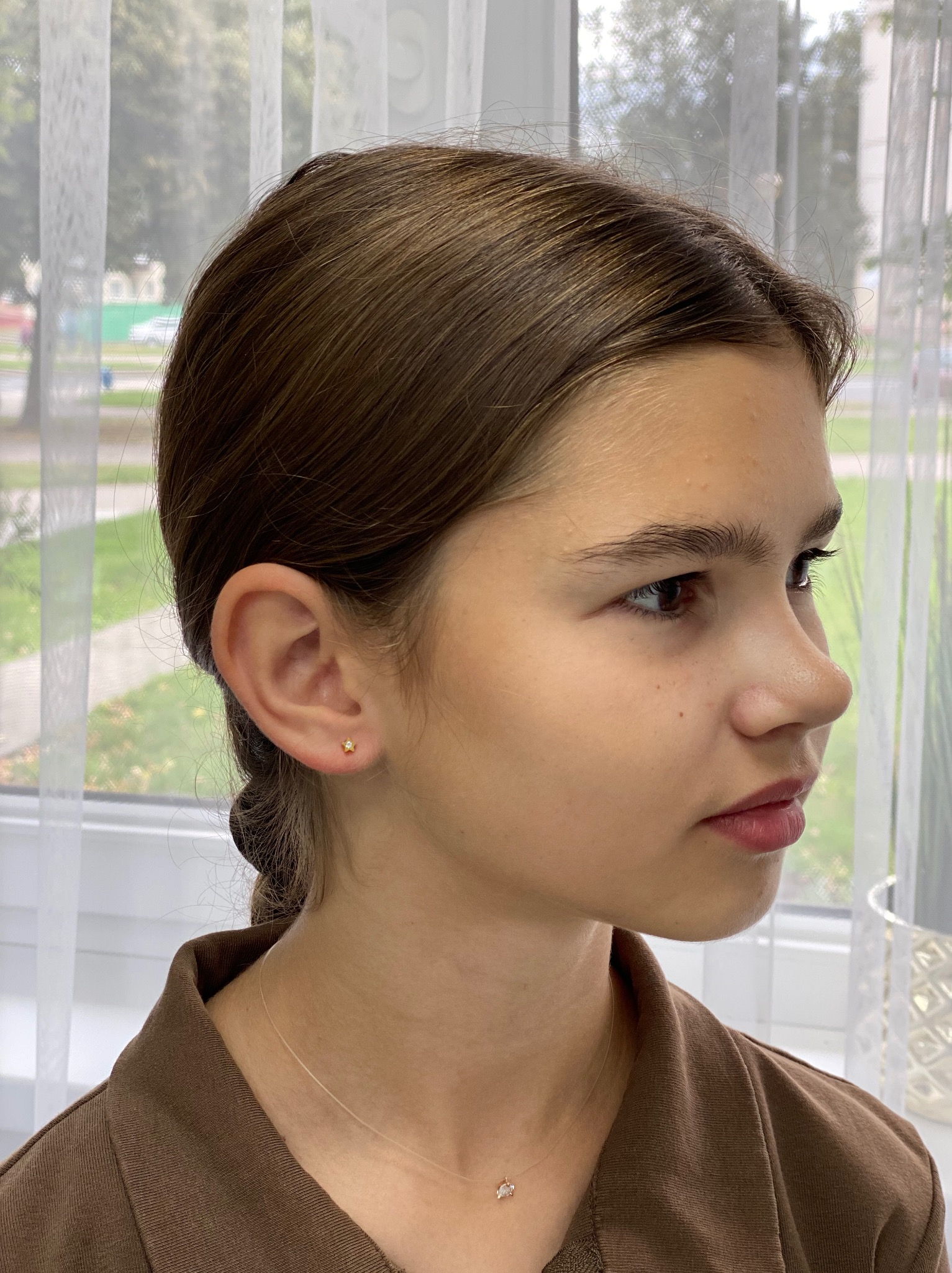 Profil młodej kobiety z kolczykiem w kształcie gwiazdki. Delikatny makijaż, brązowe włosy upięte z tyłu głowy. W tle okno z białymi zasłonami.
