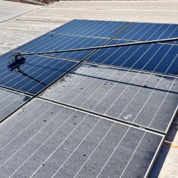 USŁUGI GASTRONOMICZNE SEBASTIAN BARTCZAK - Czyszczenie paneli fotowoltaicznych szczotką teleskopową. Część paneli umyta, widoczna różnica w czystości. Praca na wysokości, osoba w czerwonych spodenkach.