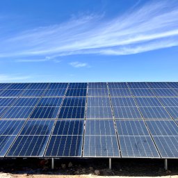 USŁUGI GASTRONOMICZNE SEBASTIAN BARTCZAK - Błękitne panele fotowoltaiczne pod błękitnym niebem z chmurami. Widoczny fragment czerwonego dachu. Instalacja na gruncie, z kurą pod spodem.