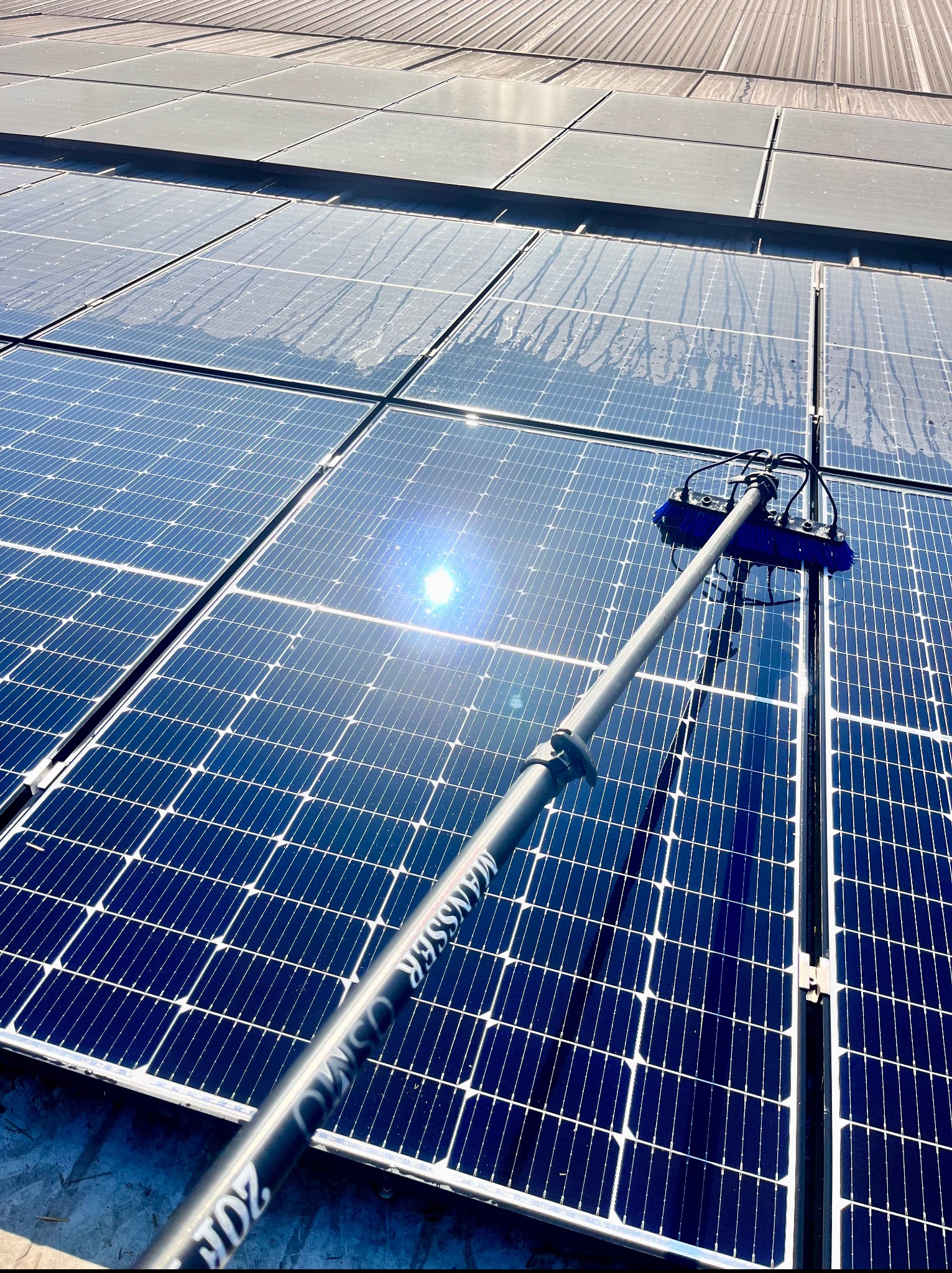 Mycie paneli fotowoltaicznych szczotką teleskopową. Widoczne ślady wody po czyszczeniu i odbicie słońca na panelach. Górny kadr pokazuje fragment dachu.