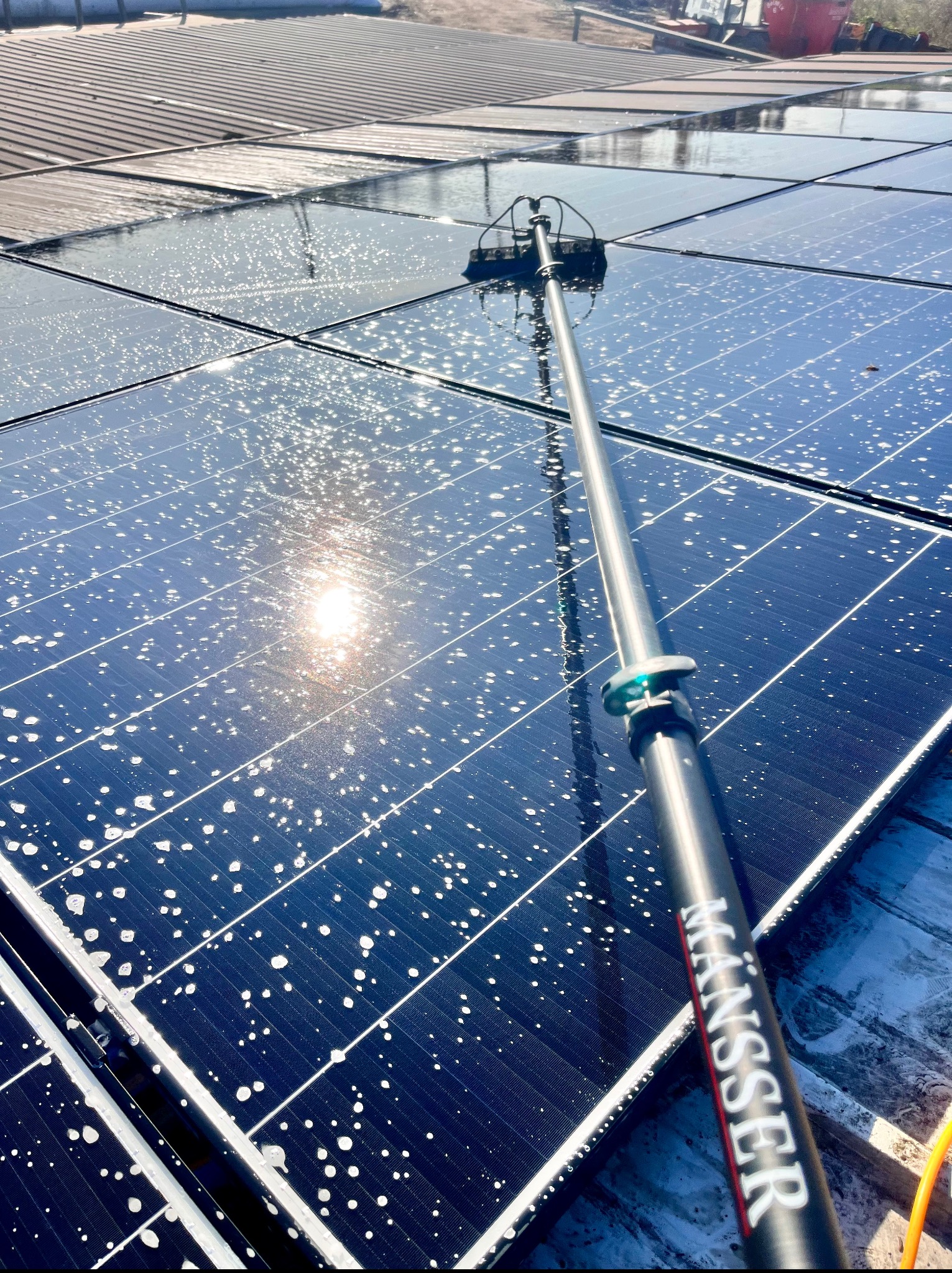 Mycie paneli fotowoltaicznych z użyciem szczotki teleskopowej. Widoczne krople wody i odbicie słońca na panelach. Czyszczenie instalacji fotowoltaicznej.