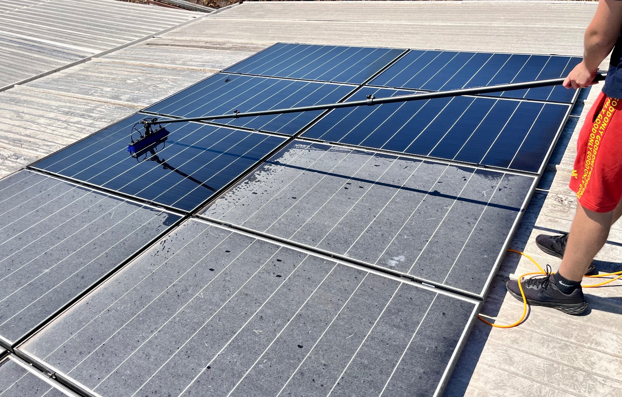 Czyszczenie paneli fotowoltaicznych szczotką teleskopową. Część paneli umyta, widoczna różnica w czystości. Praca na wysokości, osoba w czerwonych spodenkach.