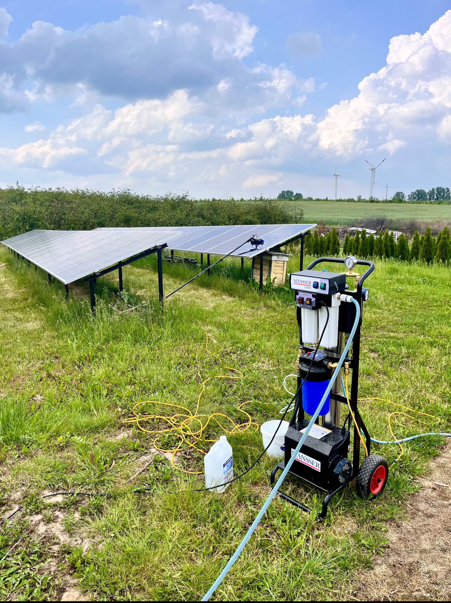 Panel słoneczny z urządzeniem czyszczącym Mansser na trawie, w tle turbiny wiatrowe i błękitne niebo. Sprzęt do mycia paneli fotowoltaicznych.