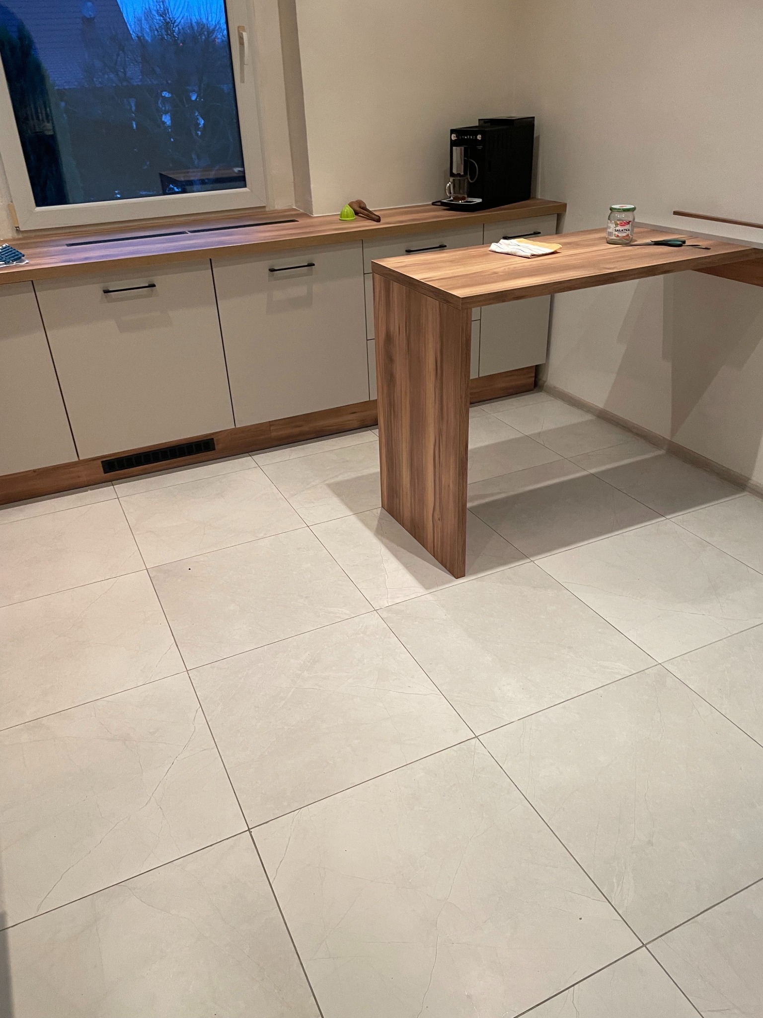 Jasne, kwadratowe płytki podłogowe w nowocześnie wykończonej kuchni z drewnianym blatem i barkiem. Widoczne okno i sprzęt AGD.