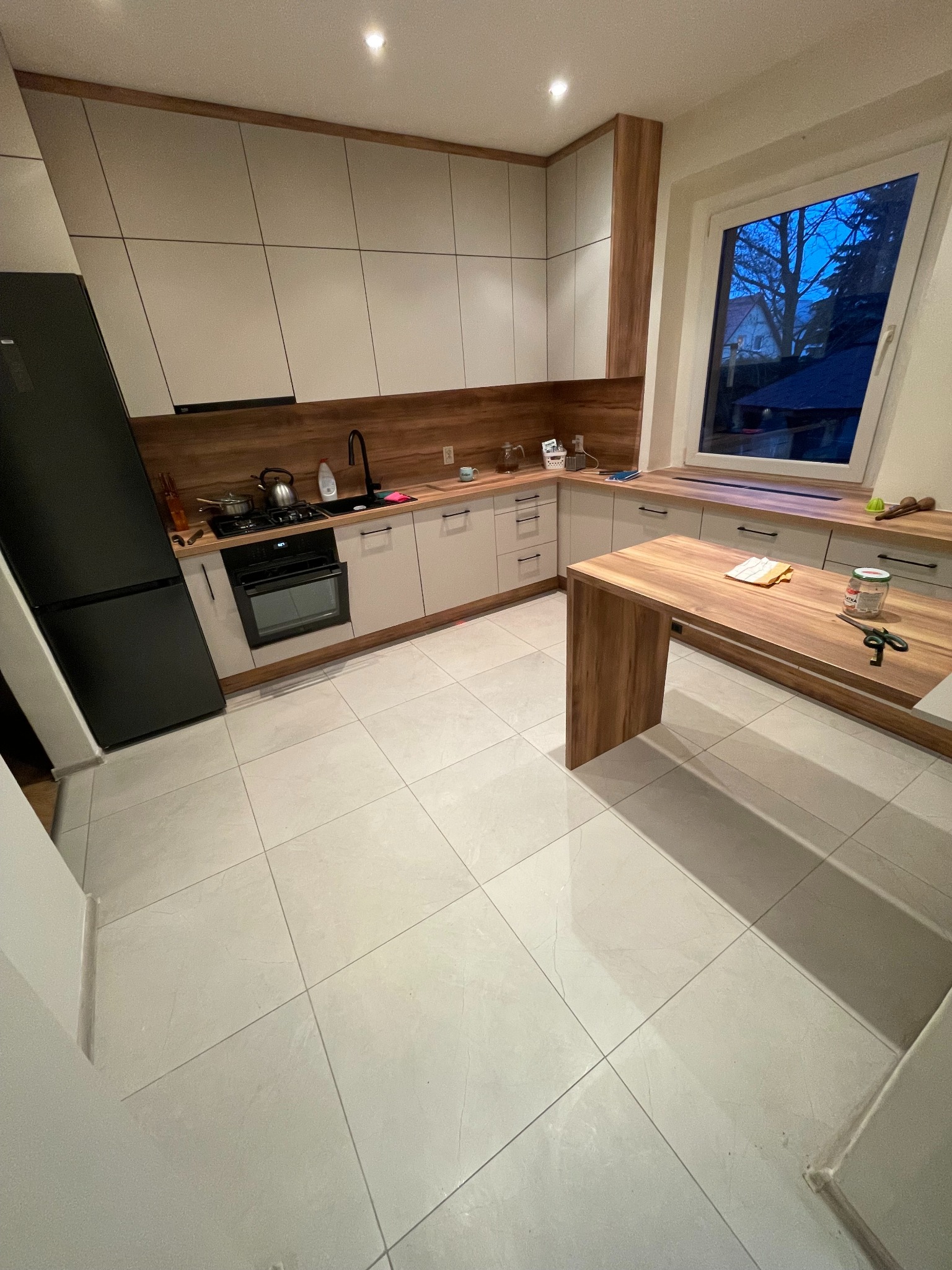 Nowoczesna kuchnia z białymi szafkami, drewnianym blatem i szarą podłogą z płytek. Widok na okno z widokiem na zewnątrz. Minimalistyczny design.