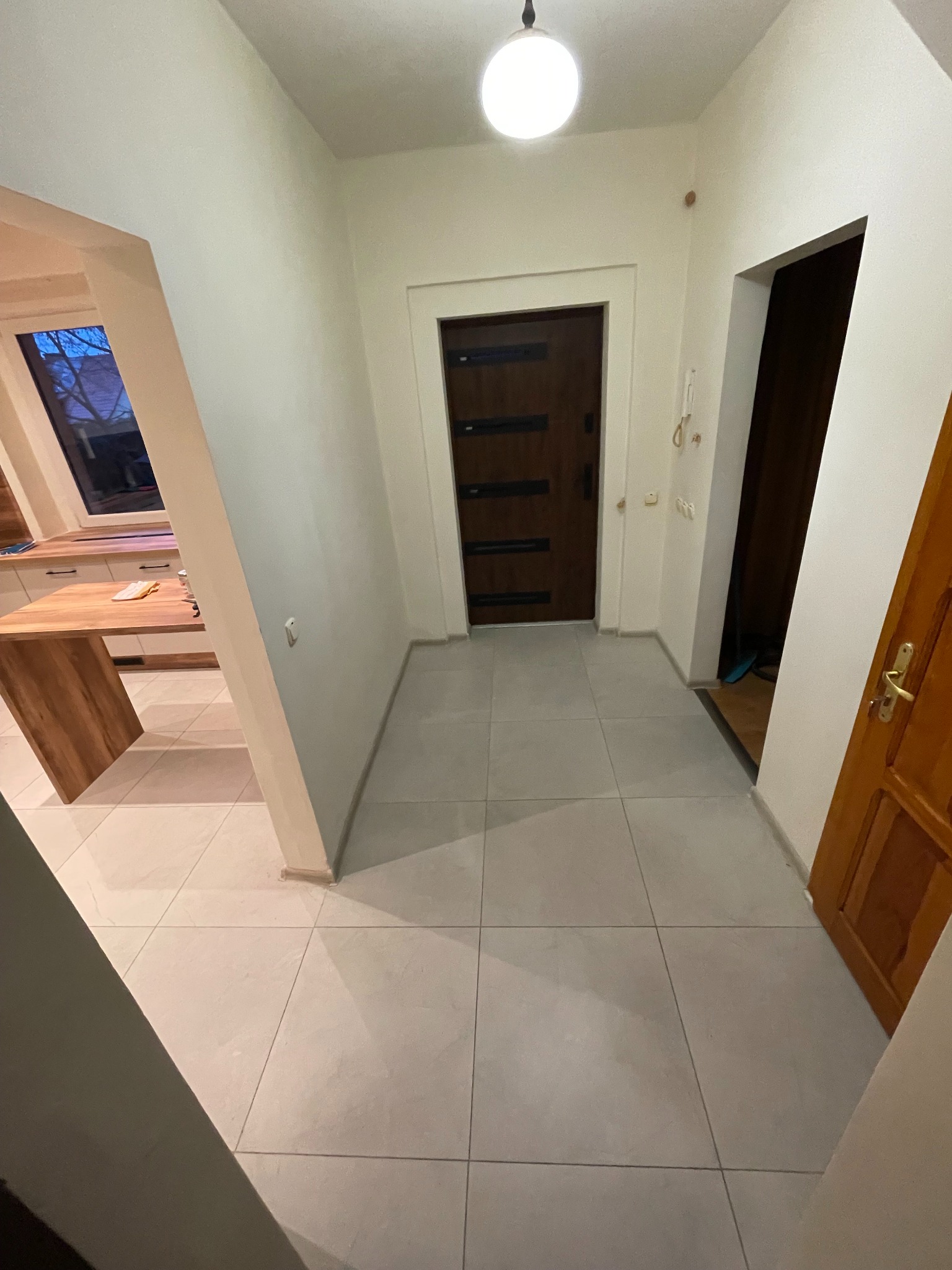 Jasne, kwadratowe płytki podłogowe w korytarzu z drewnianymi drzwiami i widokiem na kuchnię. Minimalistyczny design, neutralna kolorystyka, równomierne oświetlenie.