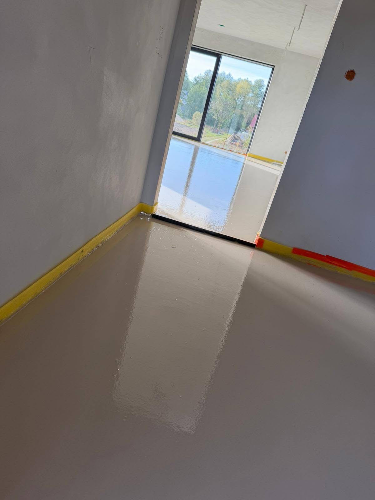 Świeżo wylana, gładka posadzka w jasnym odcieniu szarości, zabezpieczona żółtą taśmą przy ścianach. Widok na okno z zielenią na zewnątrz.