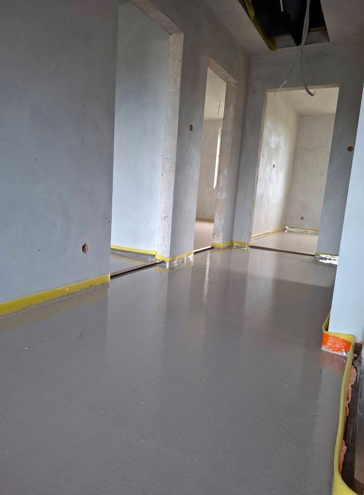 Świeżo wylana, gładka posadzka anhydrytowa w surowym wnętrzu z żółtą taśmą dylatacyjną przy ścianach i futrynach. Widoczne otwory w ścianach na instalacje.