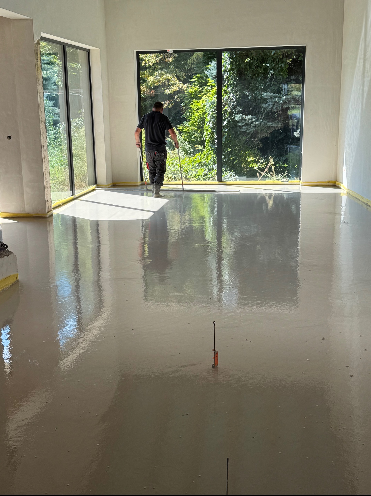 Świeżo wylana, gładka posadzka w jasnym pomieszczeniu z dużymi oknami. Pracownik wygładza powierzchnię. Widoczne narzędzia i taśmy zabezpieczające przy ścianach.