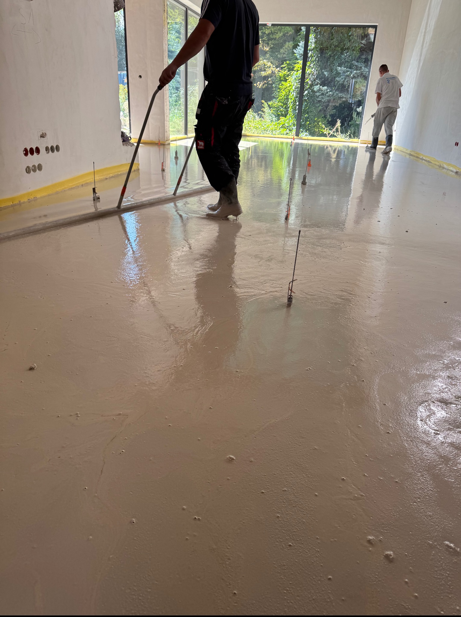 Wyrównywanie wylewki w jasnym wnętrzu z dużym oknem. Widoczne narzędzia i poziomnice. Dwóch pracowników w trakcie pracy nad posadzką. Żółta taśma zabezpieczająca ściany.