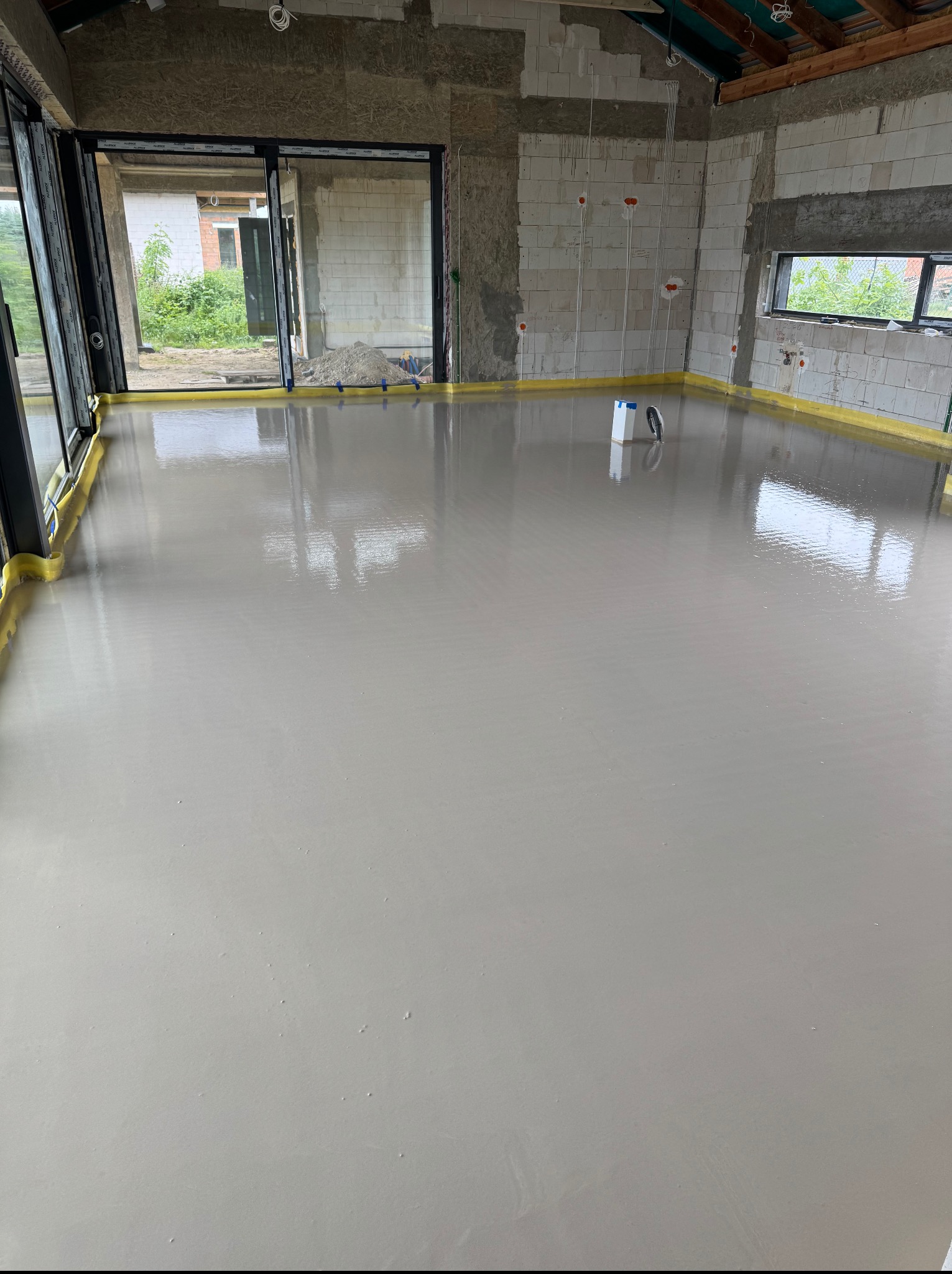 Świeżo wylana, gładka posadzka w surowym wnętrzu z widocznymi instalacjami elektrycznymi i oknami. Żółta taśma zabezpieczająca przy ścianach.