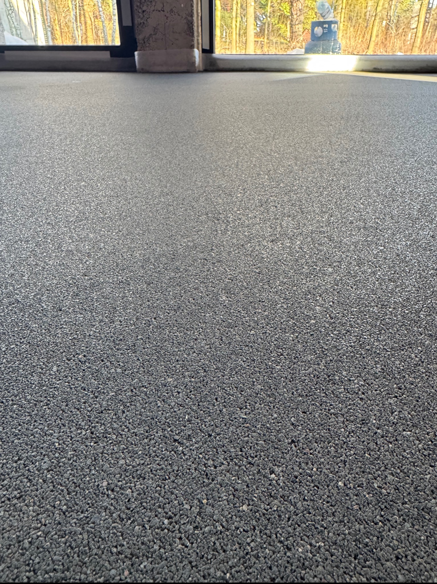 Szara, drobnoziarnista posadzka w pomieszczeniu z oknem. Widoczna faktura i równa powierzchnia. W tle zarys drzew za oknem i element konstrukcyjny budynku.