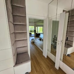 MR ATELIER SPÓŁKA Z OGRANICZONĄ ODPOWIEDZIALNOŚCIĄ - Nowoczesny przedpokój z zabudową: biała szafa z tapicerowanym siedziskiem i panelem ściennym w odcieniach szarości, lustrzane drzwi. Widok na salon.