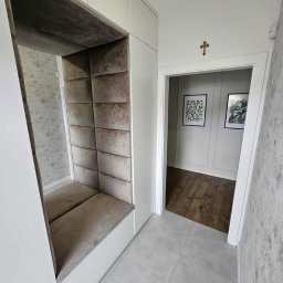 MR ATELIER SPÓŁKA Z OGRANICZONĄ ODPOWIEDZIALNOŚCIĄ - Tapicerowana wnęka z siedziskiem w zabudowie szafy w przedpokoju. Szare panele i siedzisko, białe szafy, płytki podłogowe. Widok na wnętrze przez otwarte drzwi.