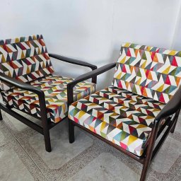 MR ATELIER SPÓŁKA Z OGRANICZONĄ ODPOWIEDZIALNOŚCIĄ - Dwa odnowione fotele z ciemnego drewna, obite tkaniną w geometryczne, kolorowe wzory, stoją na jasnej podłodze w pomieszczeniu z białą ścianą w tle.