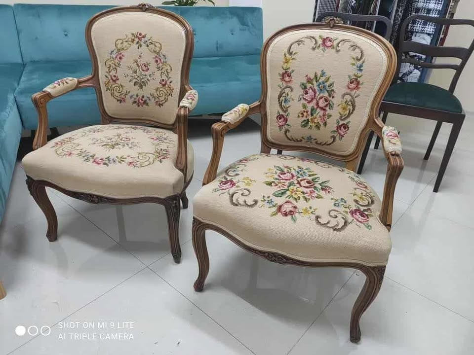 Dwa odrestaurowane, tapicerowane krzesła w stylu vintage z drewnianymi ramami i kremową tkaniną w kwiatowe wzory, na tle turkusowej sofy.