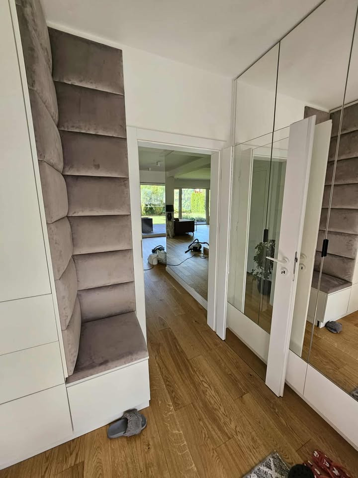 Nowoczesny przedpokój z zabudową: biała szafa z tapicerowanym siedziskiem i panelem ściennym w odcieniach szarości, lustrzane drzwi. Widok na salon.