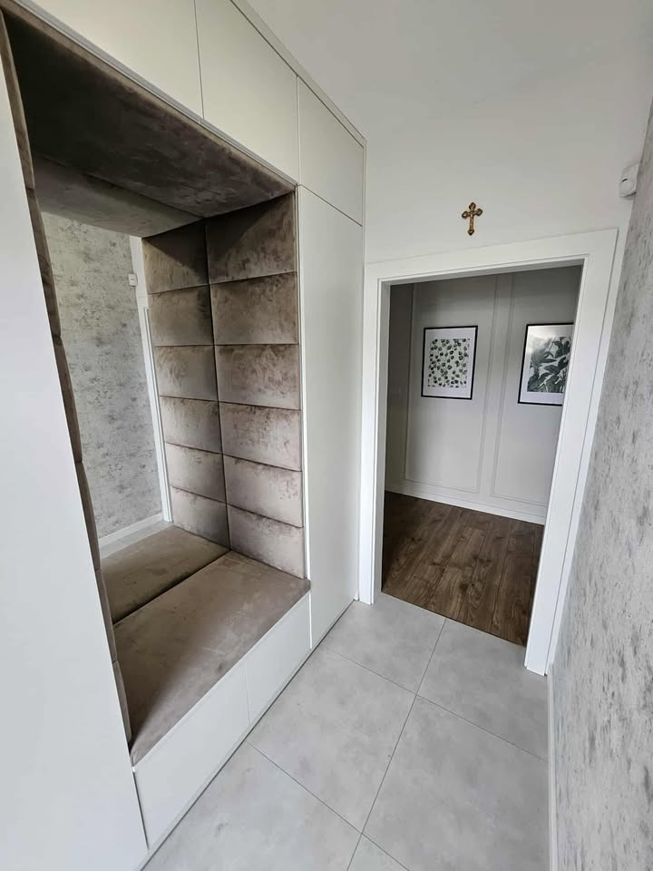 Tapicerowana wnęka z siedziskiem w zabudowie szafy w przedpokoju. Szare panele i siedzisko, białe szafy, płytki podłogowe. Widok na wnętrze przez otwarte drzwi.