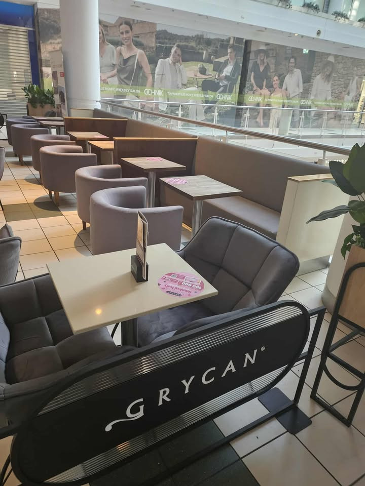 Tapicerowane fotele i kanapy w kawiarni Grycan, ustawione w rzędach. Widoczne stoliki z naklejkami promocyjnymi. W tle witryna sklepu odzieżowego Ochnik.