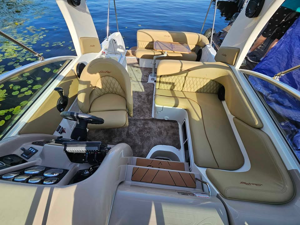 Beżowe, pikowane obicia i siedzenia w luksusowej motorówce Sea Ray, widok z kokpitu na część wypoczynkową z drewnianym stolikiem. W tle woda i roślinność.