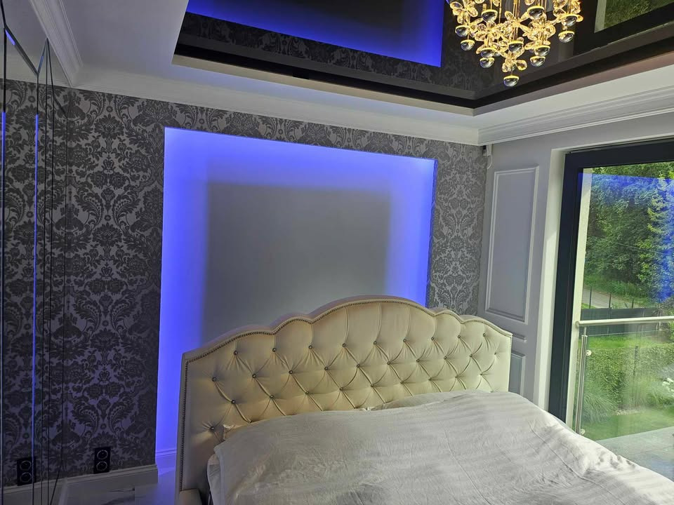 Elegancka sypialnia z tapicerowanym zagłówkiem w stylu glamour, oświetleniem LED i wzorzystą tapetą. Widok na zieleń zza okna.