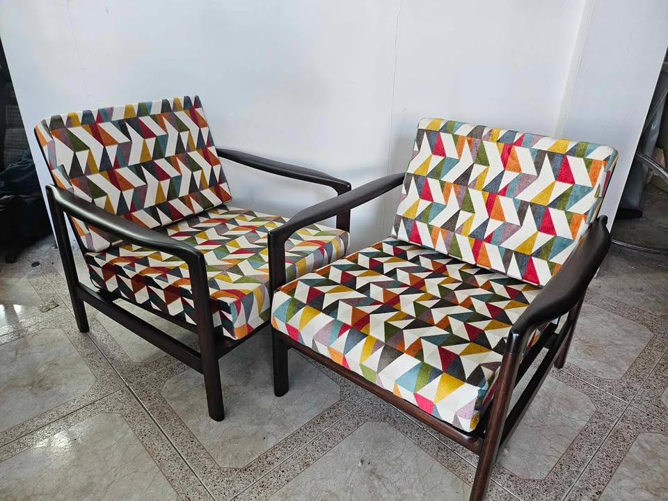 Dwa odnowione fotele z ciemnego drewna, obite tkaniną w geometryczne, kolorowe wzory, stoją na jasnej podłodze w pomieszczeniu z białą ścianą w tle.