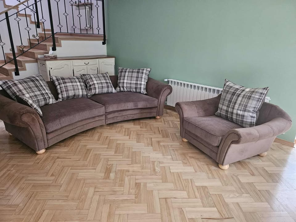 Brązowy, tapicerowany komplet wypoczynkowy: sofa o zaokrąglonym kształcie i fotel, z poduszkami w kratę, na parkiecie w jodełkę, na tle zielonej ściany.