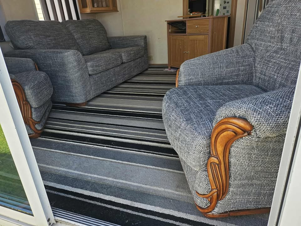 Szary, tapicerowany komplet wypoczynkowy (sofa i fotel) z drewnianymi zdobieniami na tle prugowanego dywanu i drewnianej komody w jasnym wnętrzu.