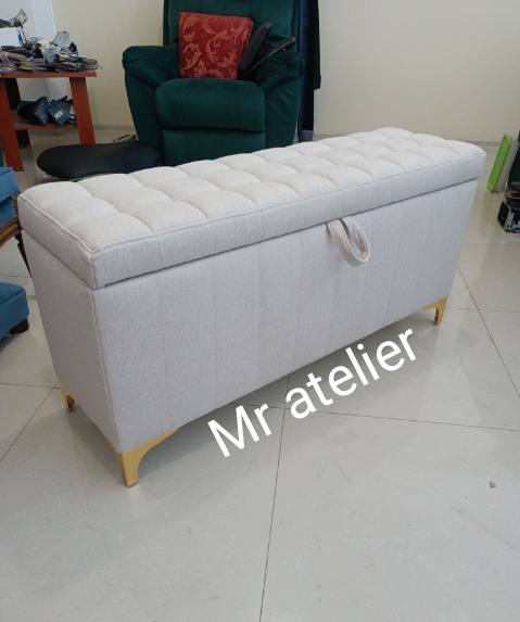 Tapicerowana skrzynia z kremową tkaniną i pikowaniem, na złotych nóżkach. W tle fotel i stolik. Widoczny uchwyt do otwierania skrzyni.