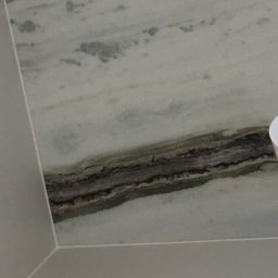 nowysilikon - Fragment ściany wykończonej płytami imitującymi marmur z wyraźnym, ciemniejszym pasmem. Widoczny narożnik i fragment sufitu. Minimalistyczny design łazienki.