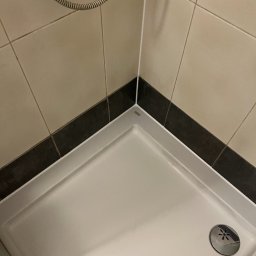 nowysilikon - Biały brodzik kabiny prysznicowej z odpływem, obłożony beżowymi płytkami z czarnym pasem. W rogu widoczny przewód prysznicowy.