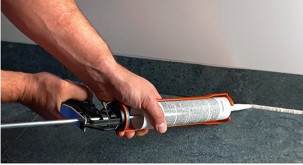 Aplikacja silikonu na styku ściany i podłogi z płytek. Widoczne dłonie, pistolet do silikonu i świeżo nałożona fuga. Ujęcie z bliska, skupione na detalu wykończeniowym.