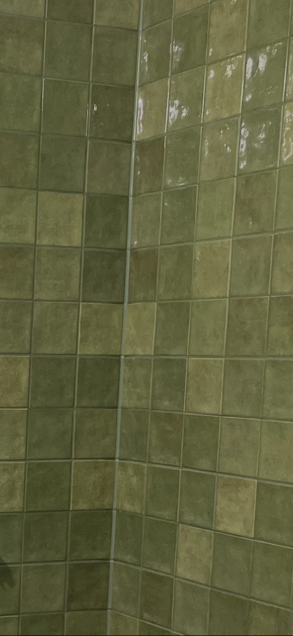 Ściana wyłożona kwadratowymi, oliwkowymi płytkami ceramicznymi z widoczną fugą i narożnikiem. Płytki mają delikatną, nieregularną fakturę i połysk.