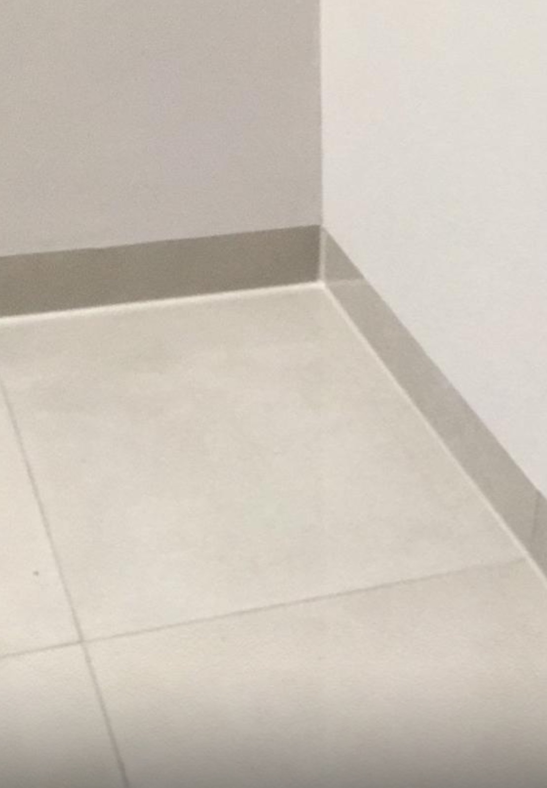 Jasne, kwadratowe płytki podłogowe z szarą listwą przypodłogową w rogu pomieszczenia. Minimalistyczny design, neutralna kolorystyka, gładka powierzchnia.