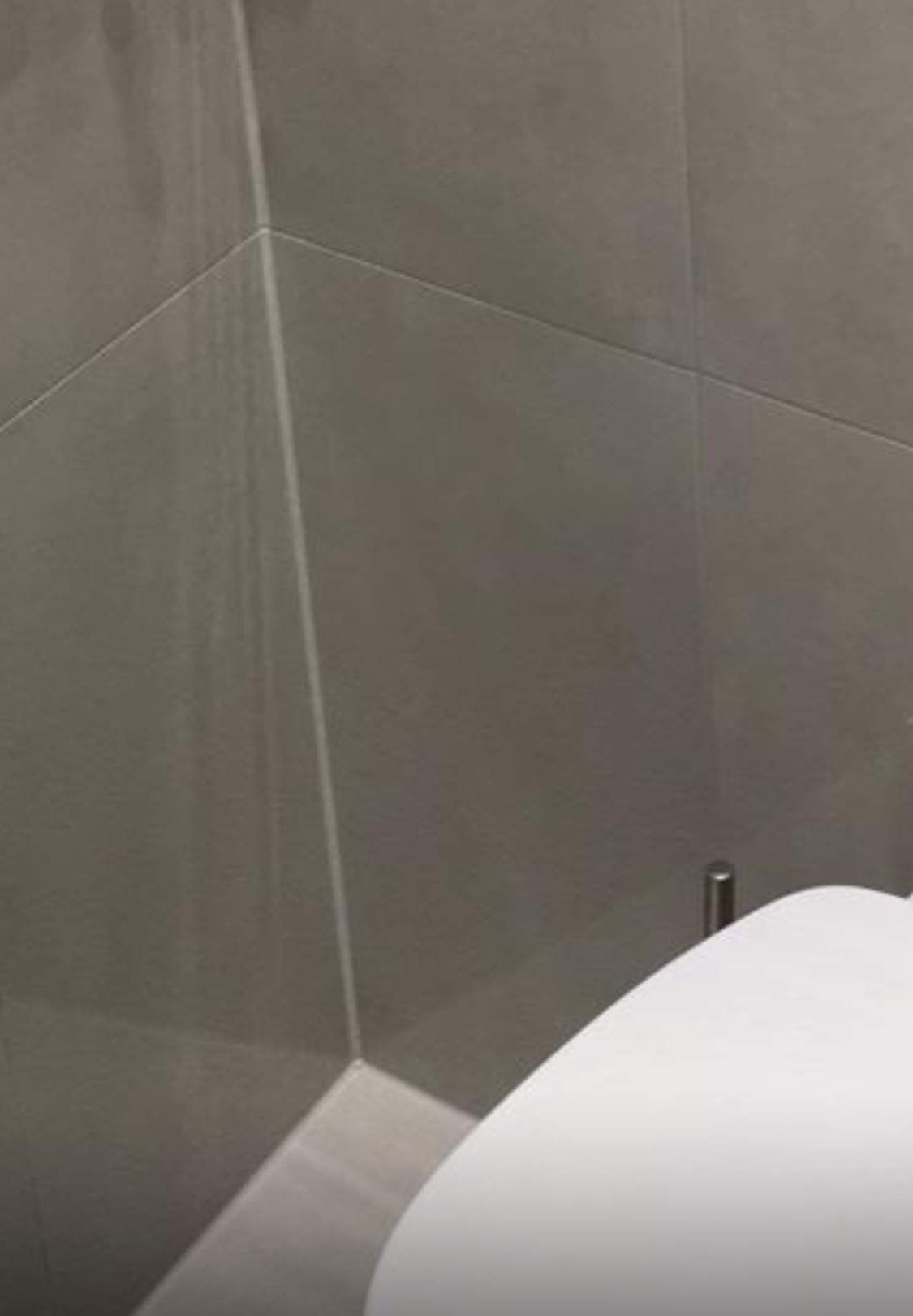 Szare, matowe płytki w rogu łazienki, z widocznym fragmentem białej umywalki. Minimalistyczne wykończenie, precyzyjne fugi, nowoczesny design.