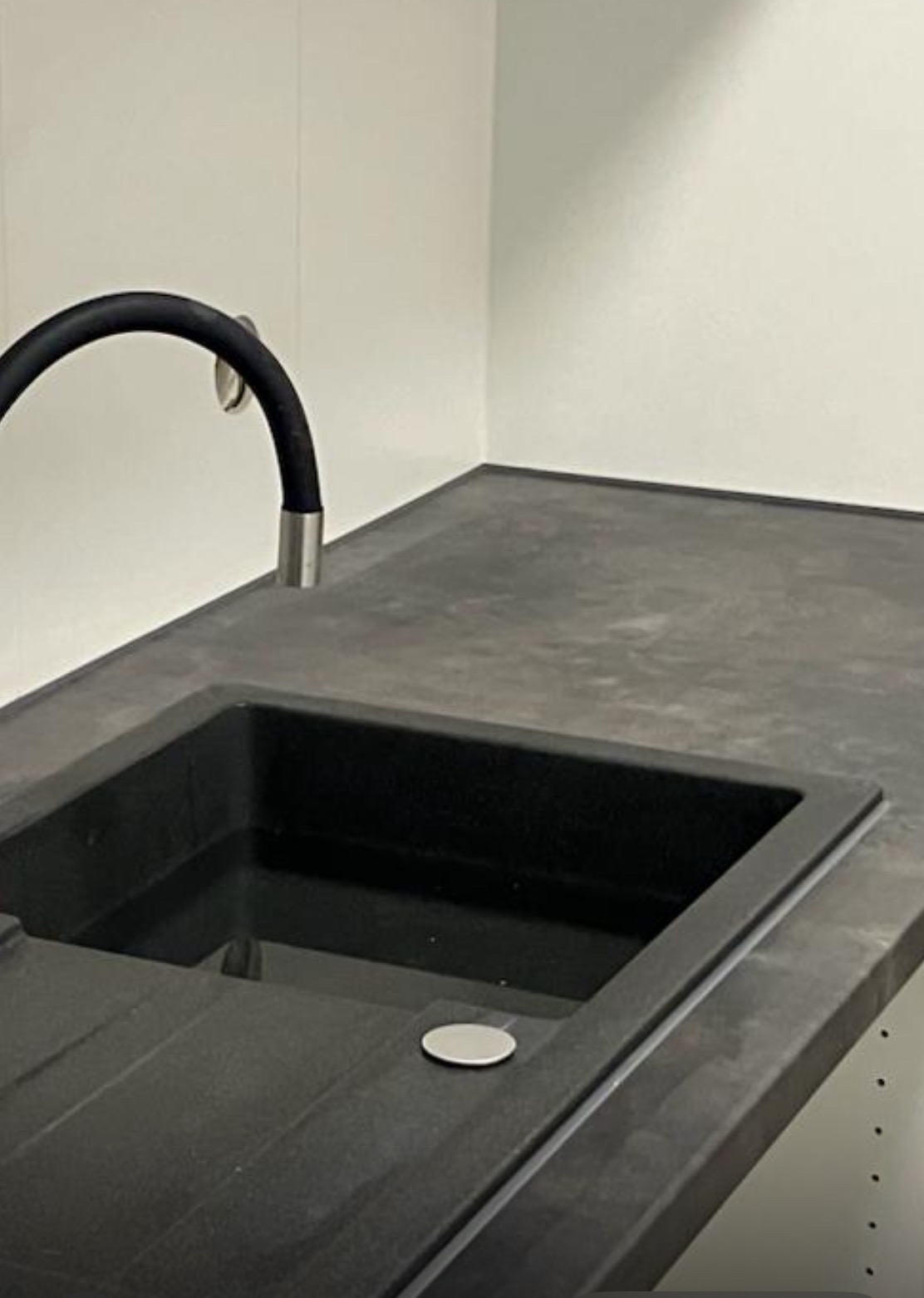 Nowoczesna, czarna umywalka z elastyczną baterią na blacie z szarego kamienia, minimalistyczne wnętrze łazienki z białymi ścianami.